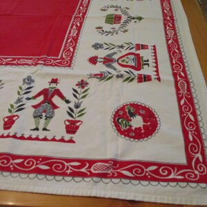 May include: A rectangular tablecloth featuring a solid red center and a white border decorated with red and green folk-art style illustrations of figures, flowers, and a circular rooster motif, framed by a red and white scrolling vine pattern.