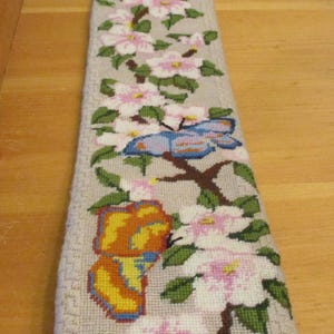 May include: Needlepoint wall hanging featuring a floral and butterfly design. The beige background is adorned with pink and white flowers, green leaves, and two colorful butterflies. The top has a decorative metal hanger.