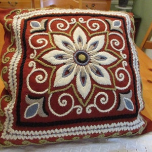 May include: A square, decorative pillow with a red background and an embroidered floral design. The flower is white, blue, and tan, with a dark center. The pillow has a white and black border and is placed on a wooden surface.