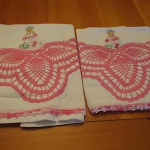 May include: Two white pillowcases with pink crocheted lace trim and a pink lace design. Each pillowcase has an embroidered doll figure in the upper corner. The pillowcases are displayed on a wooden surface.
