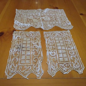 May include: Three pieces of off-white lace fabric featuring intricate openwork patterns. One larger rectangular piece is positioned horizontally above two smaller, matching rectangular pieces, all displaying detailed floral and geometric motifs.
