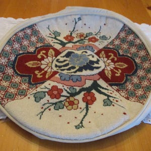 May include: Round, off-white needlepoint with a floral design. The center features a large, ornate floral motif in red, blue, and cream. The border has a geometric pattern in red, green, and cream. The piece is framed with a white, padded edge.