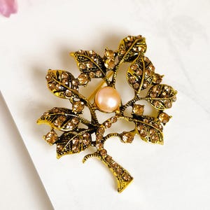 Peut inclure: Broche dorée en forme d'arbre avec une perle centrale de couleur pêche. Les feuilles et les branches sont ornées de petits cristaux ambrés scintillants. La broche est posée sur un fond blanc.
