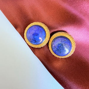 Peut inclure: Une paire de boucles d'oreilles dorées avec une bordure texturée. Chaque boucle d'oreille présente une grosse pierre ronde bleu foncé. Les boucles d'oreilles sont posées sur un fond bordeaux avec une rayure diagonale blanche et bordeaux.