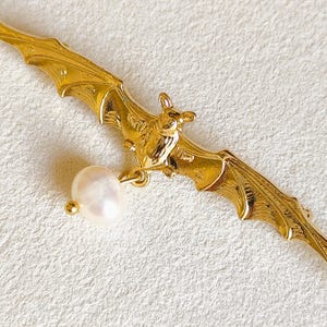 Peut inclure: Une broche en métal doré en forme de chauve-souris aux ailes déployées. La chauve-souris présente des textures d'ailes détaillées et une petite perle blanche ronde suspendue au corps par un fin maillon en métal doré, créant un contraste.