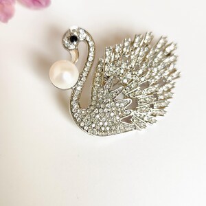 Peut inclure: Broche en forme de cygne argenté, incrustée de strass transparents. Le cygne a une perle et un œil en gemme noire. La broche est posée sur un fond blanc, avec une fleur rose floue dans le coin supérieur gauche.
