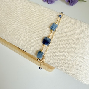 Peut inclure: Un bracelet en chaîne dorée orné de pierres bleues carrées et rondes. Le bracelet est présenté sur un tissu crème, avec des fleurs violettes en arrière-plan. Le design du bracelet comprend une série de petites perles bleues rondes et de pierres plus grandes taillées en carré.