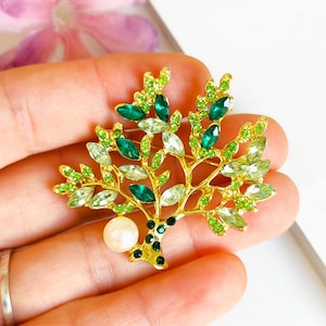 Peut inclure: Broche en forme d'arbre doré, ornée de feuilles en cristaux verts et transparents et d'un accent perlé. La broche est tenue dans une main, avec une fleur rose et un fond blanc visibles.