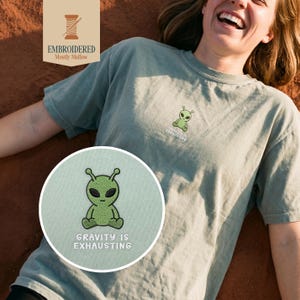 May include: Sage green t-shirt with an embroidered alien design and the text "GRAVITY IS EXHAUSTING". The shirt also features a small embroidered logo of an alien. The shirt is made of a soft, comfortable material.