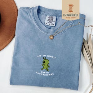 May include: A blue Comfort Colors t-shirt with the embroidered phrase "OFF TO COMMIT EARTH SHENANIGANS" and a green alien graphic. A gold necklace and a brown hat are also visible.