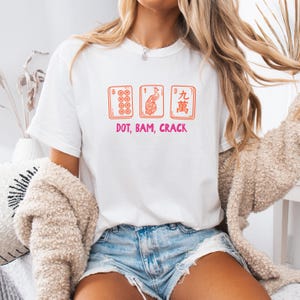 May include: White t-shirt featuring three orange Mahjong tiles with the text "DOT, BAM, CRACK" in pink. The tiles display the numbers 8, 1, and 9, along with a peacock design. The shirt is a casual, everyday wear item.