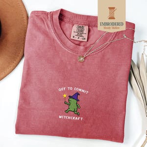 May include: A dusty rose t-shirt with the phrase "OFF TO COMMIT WITCHCRAFT" and a green frog wearing a purple witch hat and holding a wand. The shirt has a Comfort Colors tag and a silver necklace with a pendant.