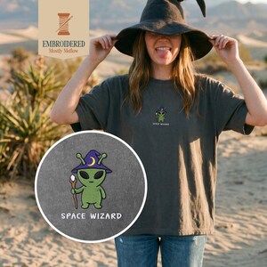 May include: Dark gray t-shirt with an embroidered green alien wearing a purple wizard hat and holding a staff. The text "SPACE WIZARD" is below the alien. The shirt also has the text "EMBROIDERED Mostly Mellow" in the upper left corner.