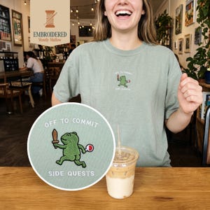 May include: Sage green t-shirt with an embroidered frog graphic holding a sword and a red ball, with the text "OFF TO COMMIT SIDE QUESTS". The shirt also features the embroidered text "EMBROIDERED Mostly Mellow".