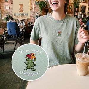 May include: Sage green t-shirt with a small embroidered frog wearing a jester's hat. The shirt has the text "OFF TO COMMIT TOMFOOLERY". The image also shows a round patch with the same design.