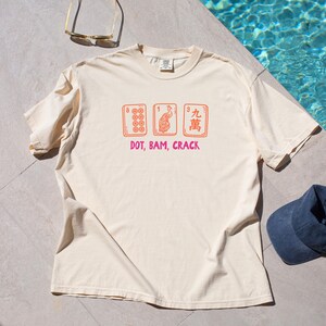 May include: Cream-colored t-shirt featuring a graphic of three Mahjong tiles in orange, with the text "DOT, BAM, CRACK" in pink. The tiles depict various symbols and numbers. A casual, comfortable tee.