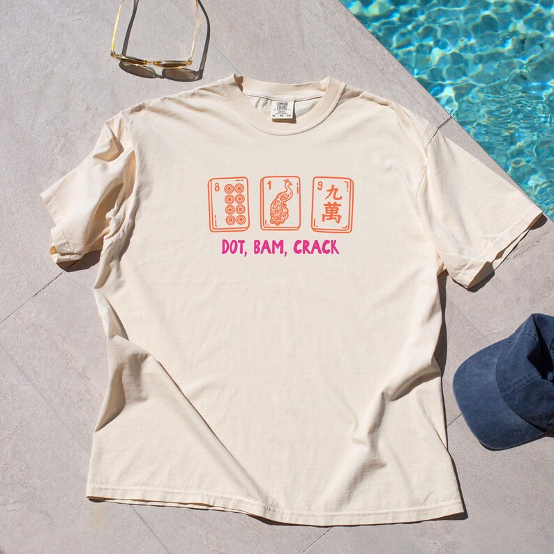 May include: Cream-colored t-shirt featuring a graphic of three Mahjong tiles in orange, with the text "DOT, BAM, CRACK" in pink. The tiles depict various symbols and numbers. A casual, comfortable tee.