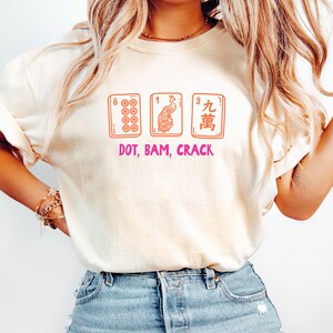 May include: Cream-colored t-shirt featuring three Mahjong tiles with the text "DOT, BAM, CRACK" in pink. The tiles are orange and depict dots, a peacock, and Chinese characters. The shirt is a casual, everyday wear item.