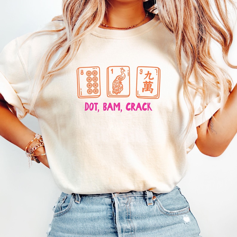 May include: Cream-colored t-shirt featuring three Mahjong tiles with the text "DOT, BAM, CRACK" in pink. The tiles are orange and depict dots, a peacock, and Chinese characters. The shirt is a casual, everyday wear item.