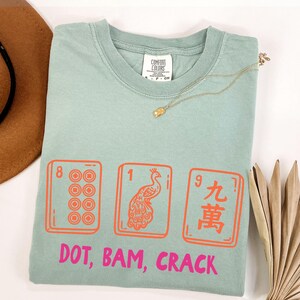 May include: A light green t-shirt with a graphic of three Mahjong tiles in orange and the text "DOT, BAM, CRACK" in pink. A gold necklace and a brown hat are visible.