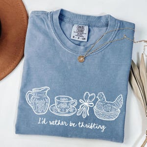 May include: A blue Comfort Colors t-shirt with a white graphic of a pitcher, teacup, spoons, and a chicken in a basket. The text "I'd rather be thrifting" is below the graphic. A gold necklace and a brown hat are also visible.