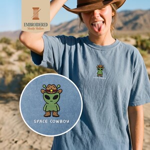 May include: A blue t-shirt with a small embroidered design of a green alien wearing a cowboy hat and boots. The text "SPACE COWBOY" is below the alien. The shirt also has the text "EMBROIDERED Mostly Mellow" in the upper left corner.