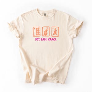 May include: Cream-colored t-shirt featuring a graphic of three Mahjong tiles in orange and red, with the text "DOT, BAM, CRACK" in pink. The shirt has short sleeves and a crew neck.