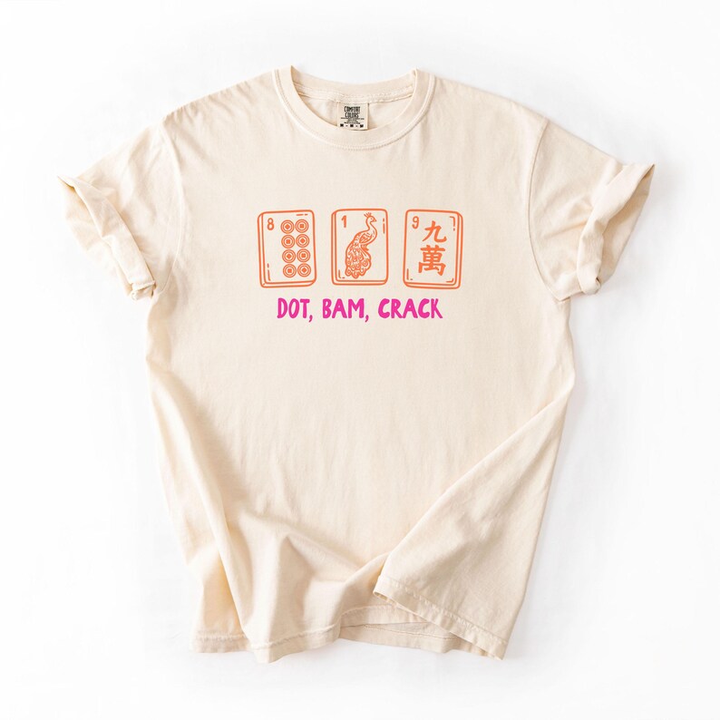 May include: Cream-colored t-shirt featuring a graphic of three Mahjong tiles in orange and red, with the text "DOT, BAM, CRACK" in pink. The shirt has short sleeves and a crew neck.