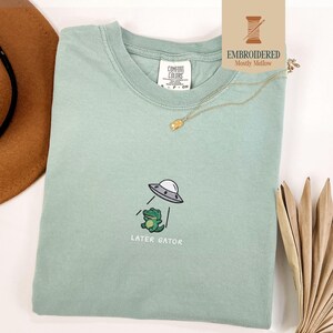 May include: A light green t-shirt with an embroidered design of a cartoon alligator being abducted by a UFO. The text "LATER GATOR" is below the design. A gold necklace and a brown hat are visible.