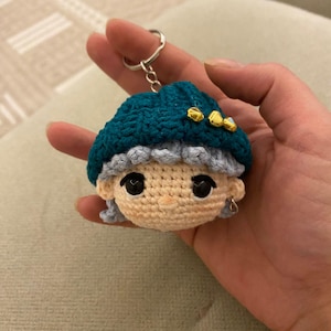 May include: Crocheted keychain featuring a doll's head with a teal hat, gray hair, and gold accents. The doll has black eyes and a silver earring. The keychain is attached to a silver ring.