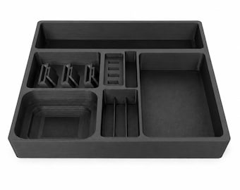 May include: A matte black rectangular desk organizer tray featuring multiple compartments of varying sizes. The layout includes a long top section, a square area with three angled slots, a small rectangular slot, a deep square bin, and two larger open sections.