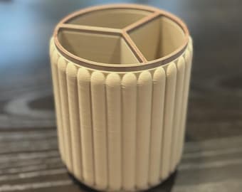 May include: A cylindrical desk organizer in a light beige color featuring a vertically ribbed exterior texture. The interior is divided into three separate compartments by internal walls, creating distinct sections for storage and organization.