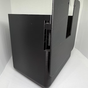 May include: A black 3D-printed plastic enclosure featuring a hinged side panel and a rectangular cutout on the front face. The structure has a smooth, matte finish with visible layer lines from the manufacturing process and a vertical hinge mechanism.