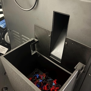 May include: A black 3D printer with an open compartment filled with colorful filament scraps. The printer has a textured finish and a sleek, modern design. A black cable and connector are visible at the top.