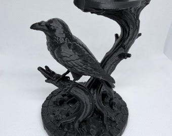 May include: A black 3D printed object featuring a raven perched on a tree branch that extends upward to form a circular holder. The base is round and textured with a cobblestone pattern, supporting the tree trunk and the bird figure.