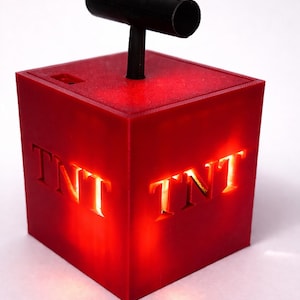 Può includere: Una luce decorativa rossa a forma di cubo con le lettere "TNT" illuminate su due lati. È dotata di una maniglia nera sulla parte superiore, che ricorda un candelotto di dinamite. La luce emette un bagliore caldo.