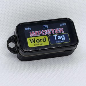 May include: A black rectangular electronic device with a screen displaying the text IMPOSTER in red, Word in yellow with sit down below it, and Tag outside in blue. The device features a small circular loop on the left side for attachment.