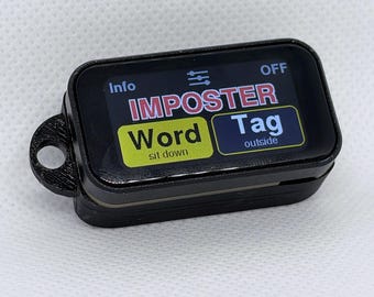 May include: A black rectangular electronic device with a screen displaying the text IMPOSTER in red, Word in yellow with sit down below it, and Tag outside in blue. The device features a small circular loop on the left side for attachment.