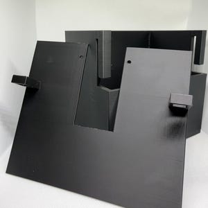 May include: A black 3D printed organizer stand featuring a flat front panel with a central cutout, two small protruding rectangular tabs on the sides, and a deeper rear section with vertical slots for holding items securely in place.