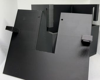 May include: A black 3D printed organizer stand featuring a flat front panel with a central cutout, two small protruding rectangular tabs on the sides, and a deeper rear section with vertical slots for holding items securely in place.