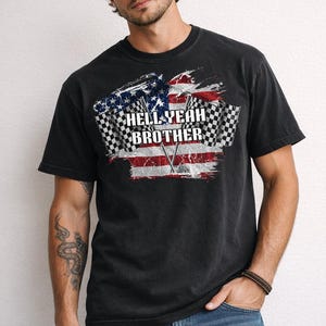 May include: Black t-shirt with a distressed American flag design and checkered racing flags. The shirt features the text "HELL YEAH BROTHER" in white. The model is wearing a brown cap and blue jeans.