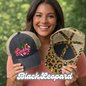 May include: A charcoal gray baseball cap with a tan mesh back and a graphic featuring a palm tree, seashell, and the word Beachy in pink neon-style text, alongside a second cap with a tan and black leopard print pattern and mesh back.