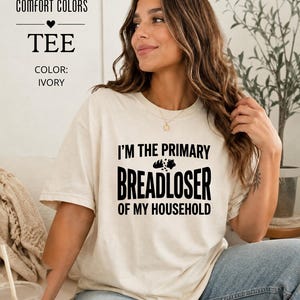 May include: An ivory short-sleeve t-shirt with black text that reads I'M THE PRIMARY BREADLOSER OF MY HOUSEHOLD, featuring a small graphic of a loaf of bread between the words PRIMARY and BREADLOSER. The shirt is a Comfort Colors brand tee.
