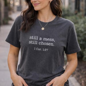 Könnte beinhalten: Dunkelgraues T-Shirt mit dem weißen Text "still a mess, still chosen." und dem Bibelvers 1 Kor. 1:27. Das T-Shirt hat einen Rundhalsausschnitt und wird getragen.