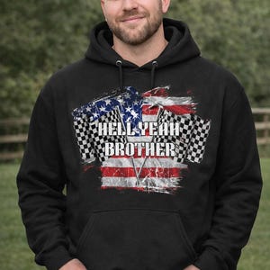 May include: Black hooded sweatshirt with a distressed American flag design and the words "HELL YEAH BROTHER". The design includes checkered racing flags. The sweatshirt is worn by a person.