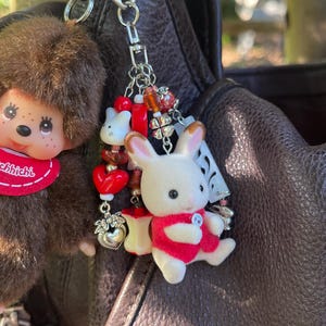 May include: A metal keychain featuring a brown fuzzy monkey doll with a red bib labeled Monchhichi, paired with a white flocked rabbit figurine wearing a red vest. The charm includes red heart beads, a silver heart charm, and a four-leaf clover.