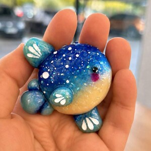 Può includere: Una piccola statuetta artigianale arrotondata a forma di pesce palla. Il corpo presenta una sezione superiore blu intenso con macchie bianche e glitter iridescenti, che sfuma verso una parte inferiore giallo pallido. Ha pinne color ottanio con segni bianchi.