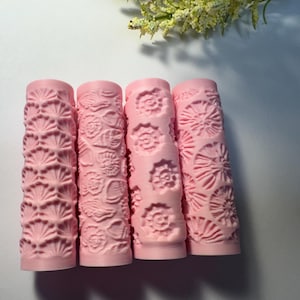May include: Four pink cylindrical rollers with embossed floral and shell patterns. The rollers are arranged vertically, showcasing detailed designs. The background is a light, neutral color, with a hint of greenery at the top.