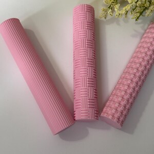 May include: Three pink textured rollers. One roller has vertical ridges, another has a woven pattern, and the third has a diamond pattern. The rollers are cylindrical and appear to be made of a soft material.
