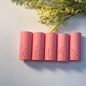 May include: Five pink cylindrical objects with floral designs are arranged horizontally. The objects are decorated with embossed floral patterns at the bottom. Yellow flowers are in the background.